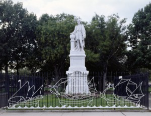 A color photograph of a white statue of Christopher Columbus standing on a base. The statue is surrounded by a black fence and and grass, with trees in the background. On the fence are the white outlines of three sailing ships. 