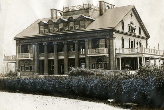 A sepia-tone photograph of a two story building surrounded by fields and bushes. The building is rectangular, with a peaked roof, pillars near the middle of the building, and a balcony on the end of the building.