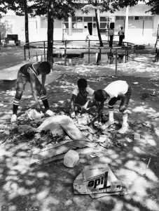 A black and white image of three children picking through a pile of trash in the middle of a playground. the side of an apartment building and playground equipment is in the background of the image.