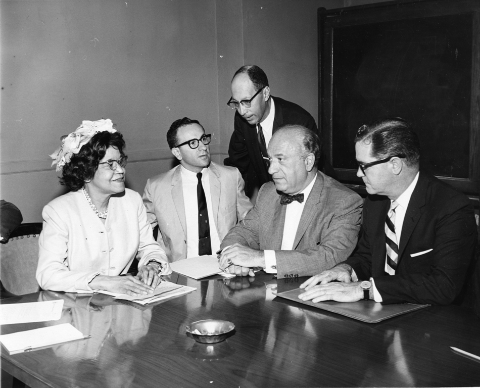 An image of a groupd o five people sitting around a desk, looking at paperwork. There are four men with suits, and one woman in a dress.