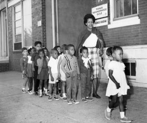 A black and white photograph of a woman walking with a group of children down a sidewalk, outside of a brick building. 