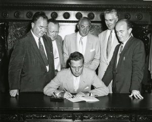 A black and white photograph of a man signing a document at a desk with a group of five men standing behind the signer. 