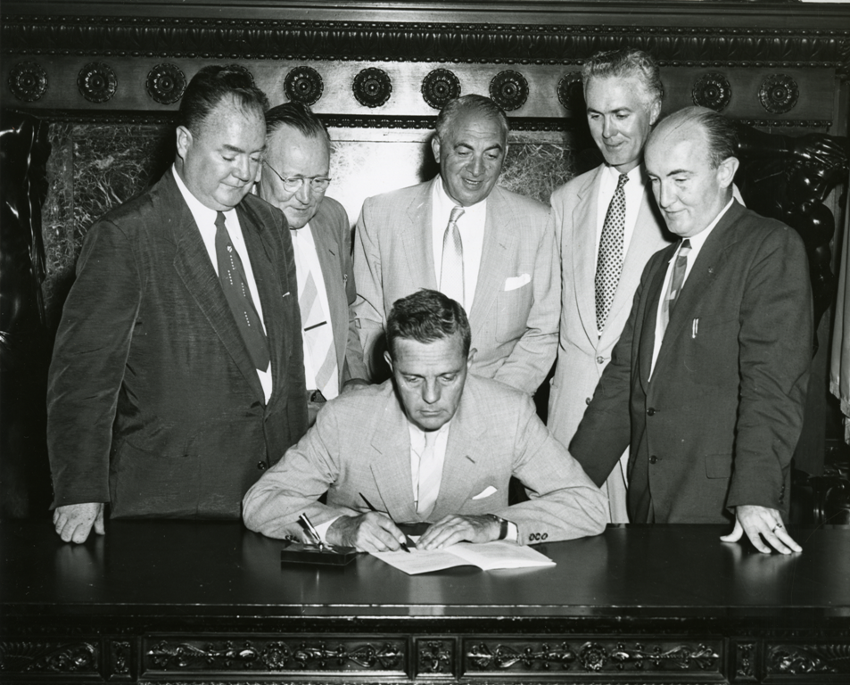 A black and white photograph of a man signing a document at a desk with a group of five men standing behind the signer.