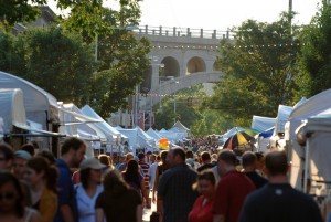 A color photograph of street vendors and artists in tents along the sidewalks of a street. A large crowd of people are walking in the middle of the street. In the background is a stone bridge. 