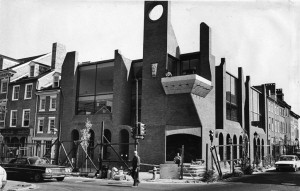 A black and white photograph of a brick building with architectural designs that are not found in the adjacent row houses There is construction equipment outside like ladders and scaffolding in front of the modern building. 