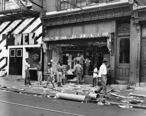 A black and white photograph of a group of people (mostly younger people) standing next to a vandalized storefront. Pieces of debris are covering the ground of the store. 