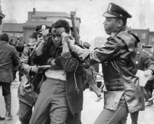 A black and white photograph of a man with blood on his face and hands by his head surrounded by two police officers holding batons. 