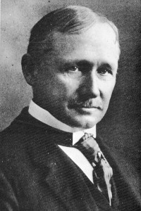 A black and white photograph a man from the shoulders up. The man is wearing a suit with a tie and is facing slightly to the right. 