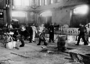 A black and white photograph of a group of men working around large pieces of machine equipment. Some men are holding tools and looking at the viewer. 