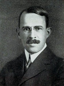 A black and white photograph of a man from the shoulders up, in a suit with glasses and a mustache looking directly at the viewer. 