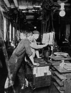 A black and white photograph of two workers in a cluttered factory looking at a time sheet. 