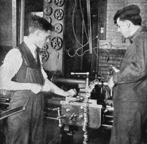 A black and white photograph of two men in overalls looking at a machine in a production shop. The man on the left of the image is holding a stopwatch. 