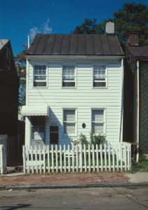 A color photograph of a two story white house, with a small white fence in front of it. Parts of the sidewalk and street are shown in front of the property.