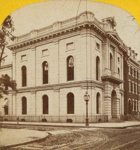 A black and white photograph showing a two-story stone building on the corner of a city block. Sections of the sidewalk and the street are visible.