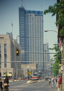 A color photograph of the PSFS Building from a few blocks away, showing the whole building, market street, people, cars, and buildings leading up to the building. 