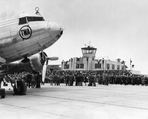 A black and white photograph of the exterior of an airport, with a large crowd of people and an airplane in the foreground. 