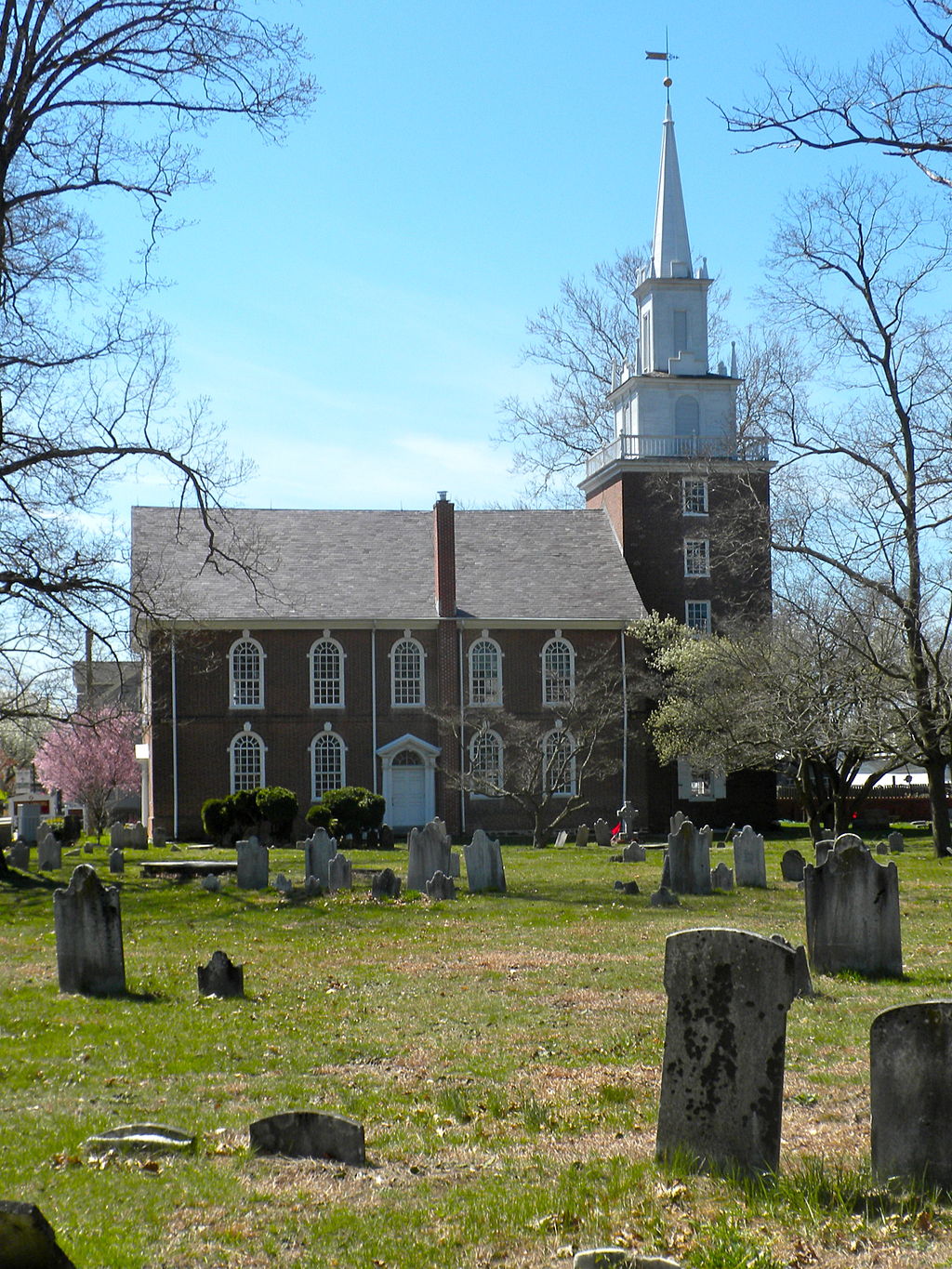 A color photograph of a church from the side. A graveyard with visible gravestones, trees, and grass is in the foreground, and the church is in the background.
