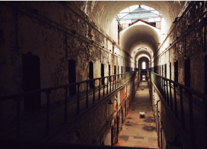 A photograph taken from the second story of cell block seven, looking down the hall to the exterior door. The cell block is in a deteriorated state but arched ceilings and glass skylights give the impression of a church or other spiritual building