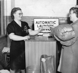 a photograph of two women using the automatic laundry service. the woman ont he left is taking a ticket from the woman on the right, who seems to be either picking up or returning her laundry