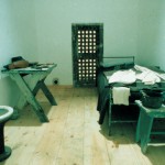 photograph of a solitary cell recreation. there is a twin bed to the right of the picture and a small night table at the foot of the bed. the left side of the room has small work table and a toilet. a door in the back of the cell leads to the work yard.