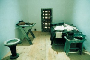 photograph of a solitary cell recreation. there is a twin bed to the right of the picture and a small night table at the foot of the bed. the left side of the room has small work table and a toilet. a door in the back of the cell leads to the work yard.