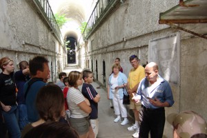 a tour guide, to the right of the photo, leads a group of visitors through a cell block, while explainging the history of eastern state to them