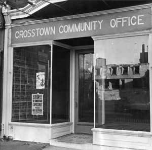 photograph of the crosstown community office storefront. In the window's reflection, delapitated row homes are visible