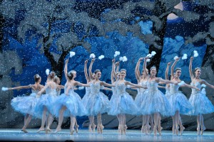 A color photograph of ballet dancers performing in The Nutcracker, as seen from the audience. The tweleve female dances on stage all wear pale blue leotards and tutus and are on point, with one arm extending over their heads as they dance in a line