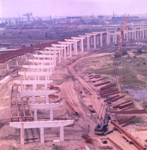 A color photograph of construction of I-95, showing pylons in place and cranes preparing to lift the road deck.