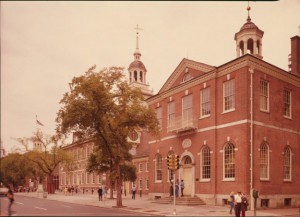 color photograph of independence hall, taken from across the street, caddy-corner to the building. a tree stands outside the entrace and a few people populate the street