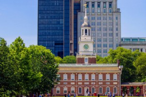 color photograph of Independence hall exterior, taken from the direct front of the building from approximately 100 feet away. Lush green trees surround the brick building and skyscrapers rise in the background against a blue sky.