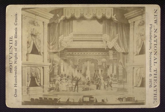 Photograph of a trade card, which has a full stage drawing of the New National Theater as it stages a play. Two levels of balconies flank the stage on either side.