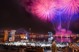 A photograph of Penn's Landing at night time. The winter festival, Winterfest, is taking place and the ice skating rink with many skaters is visible in the forground of the picture. Above, the Ben Franklin Bridge, Delaware River, and neighboring Camden are illuminated by pink and purple fireworks