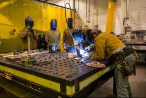 color photo shows three people in welding helmets looking on as two others in welding gear lean into a welding project atop at welding table at NextFab co-working space.