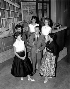 a black and white photograph of Dick Clark on the set of American Bandstand with five teenage guests