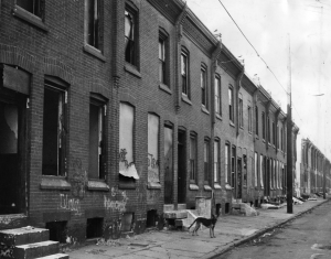 A black and while photograph of a street of row houses, many of which look abandoned or have been damaged in some way. There is trash in front of the buildings, and a dog on the sidewalk.