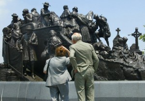 Color photograph of the Irish Memorial statue. The statue is about thirty feet wide and has many figures carved in to its bronze surface. An older couple, a man and woman, are looking at the statue with their backs to the camera.