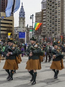 Color photograph of a group of men playing bagpipes. They are wearing kilts and other traditional Irish clothing. Behind them city hall and a large crowd of spectators is visible.