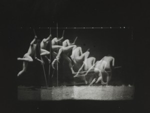 a black and white time-lapse image of a nude male pole-vaulting. It captures the figure at timed intervals, resulting in about eight apparent semi-transparent overlapping figures going through the singular motion of pole-vaulting.