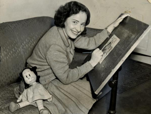 A photograph of Marjorie Buell working on an illustration while sitting at her drawing table. A 