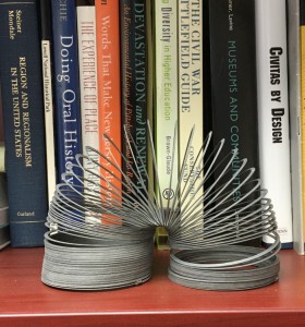 An image of a slinky in the foreground with books on a bookshelf in the background.