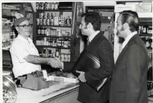 A black and white photograph of two men in business suits shaking hands with a merchant inside of a store