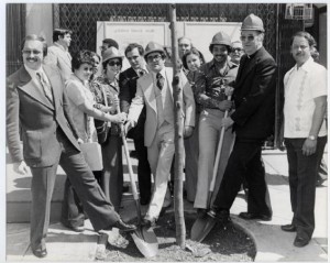 A black and white photograph of a group of people posing with a newly-planted tree and shovels