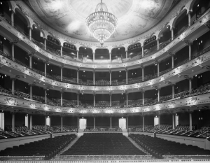 A black and white photograph of the Academy of Music from the stage, viewing the main floor seating, the three balcony levels and the large central chandelier.