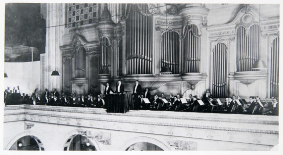 A black and white photograph of the Philadelphia Orchestra (and all members in formal attire) on the second level of the Wannamaker Store's Grand Court; behind them, Wannamaker's Great Organ.