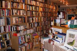 An image of the interior of Joseph Fox Bookshop featuring a wall covered in bookshelves and colorful books, with the front desk on the right-hand side (featuring more books).