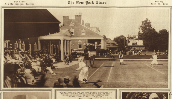 An image from the New York Times published in July 1915 of a women's doubles tennis match taken from behind the court, picturing the clubhouse and its columns to the left, along with a group of spectators under an awning.