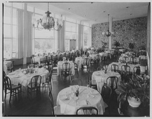 A black and white photograph of the interior of the Philadelphia Country Club dining room with windows along the left wall, two chandeliers and neatly-set tables arranged around the room. 