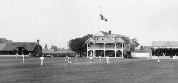 A black and white photograph of the Philadelphia Cricket Club's three-story clubhouse, with grounds in the foregrounds featuring members playing a game of cricket. A large banner is strung up to the right of the clubhouse and a crowd can be seen in the distance between the clubhouse and a steep-roofed house at the left edge of the frame.