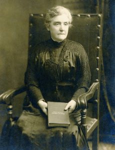 Black and white photograph of a woman seated in wooden arm chair holding a book.